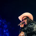 Burna Boy performing on stage in a wide-brimmed hat holding a microphone