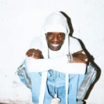 Seyi Vibez smiling in a white hood against a white brick wall