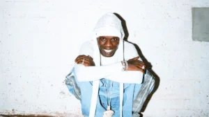 Seyi Vibez smiling in a white hood against a white brick wall