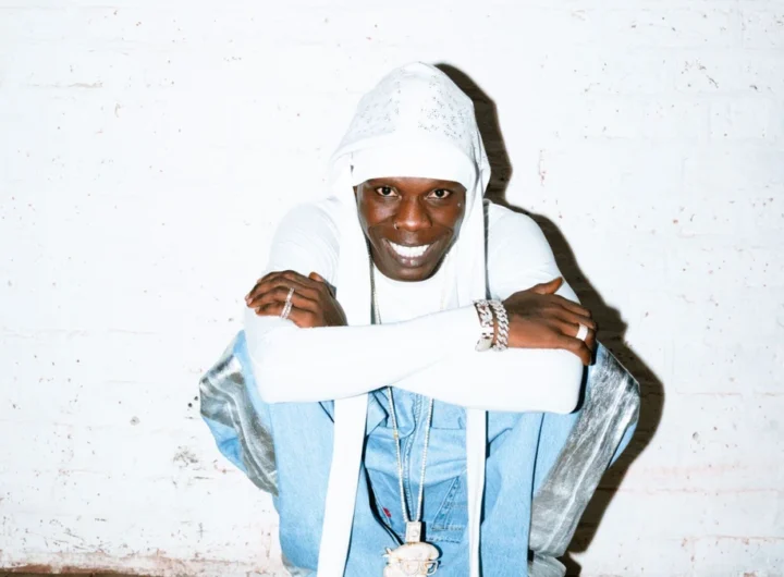 Seyi Vibez smiling in a white hood against a white brick wall