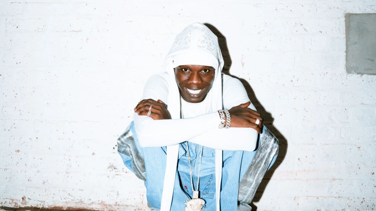 Seyi Vibez smiling in a white hood against a white brick wall