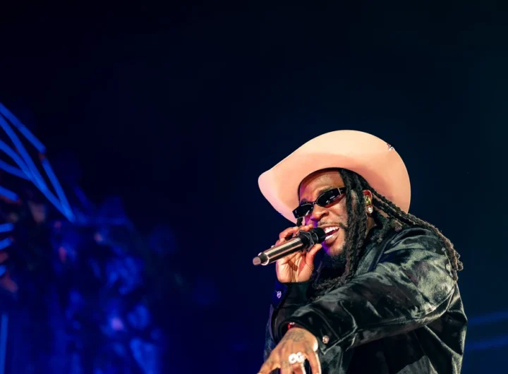 Burna Boy performing on stage in a wide-brimmed hat holding a microphone